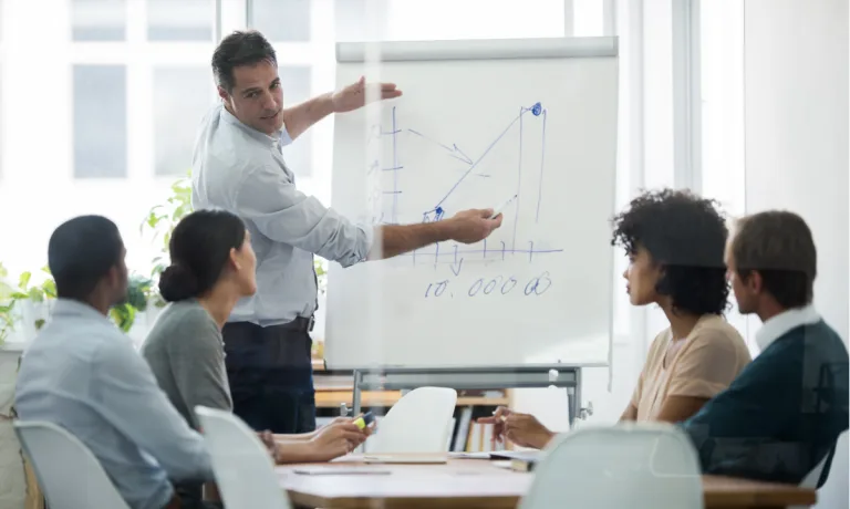 A business professional is presenting a growing graph on a flip chart to a group of attentive colleagues in a meeting room.